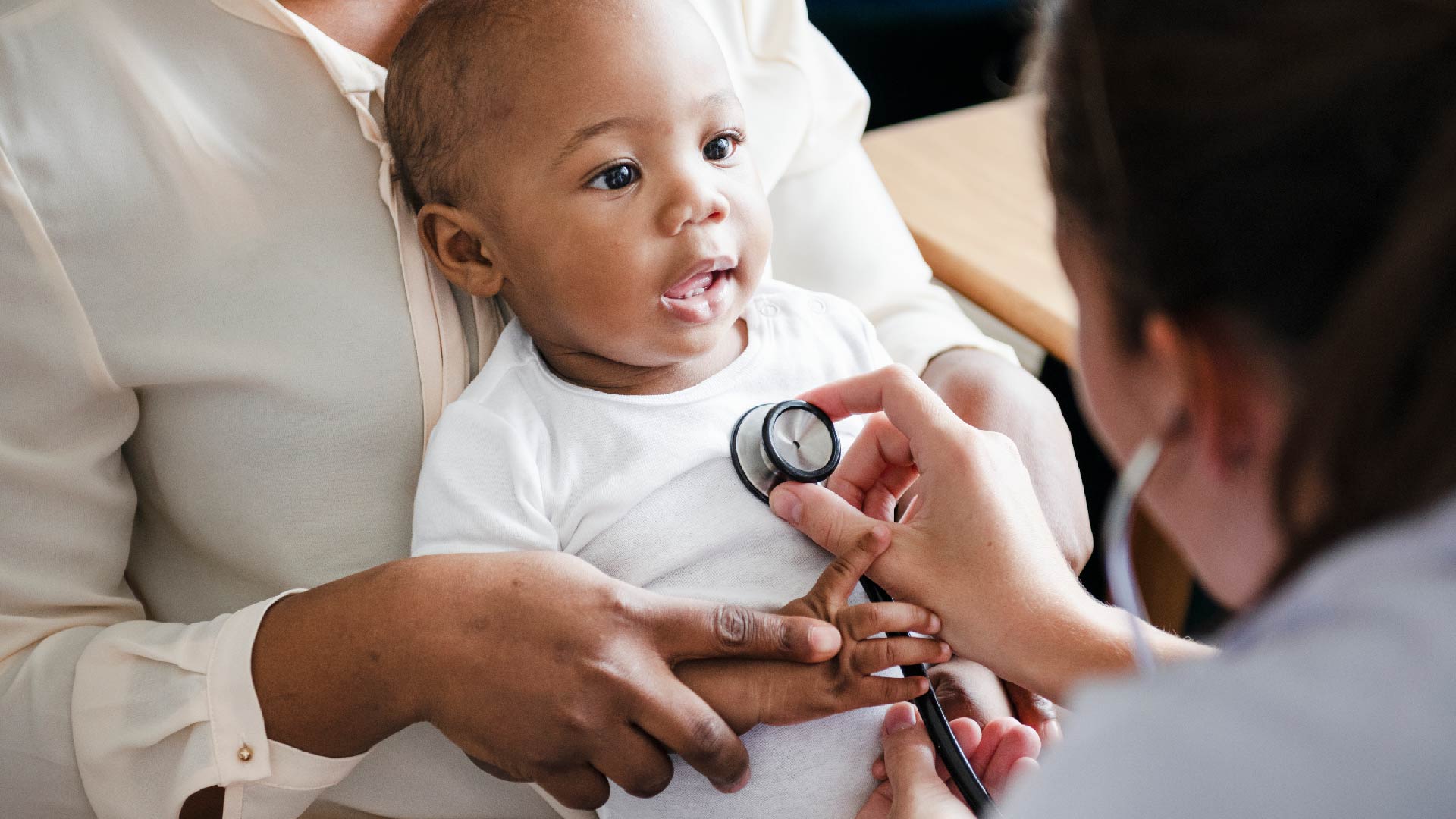 Een baby zit op de schoot van zijn mama tijdens een onderzoek bij de kinderarts