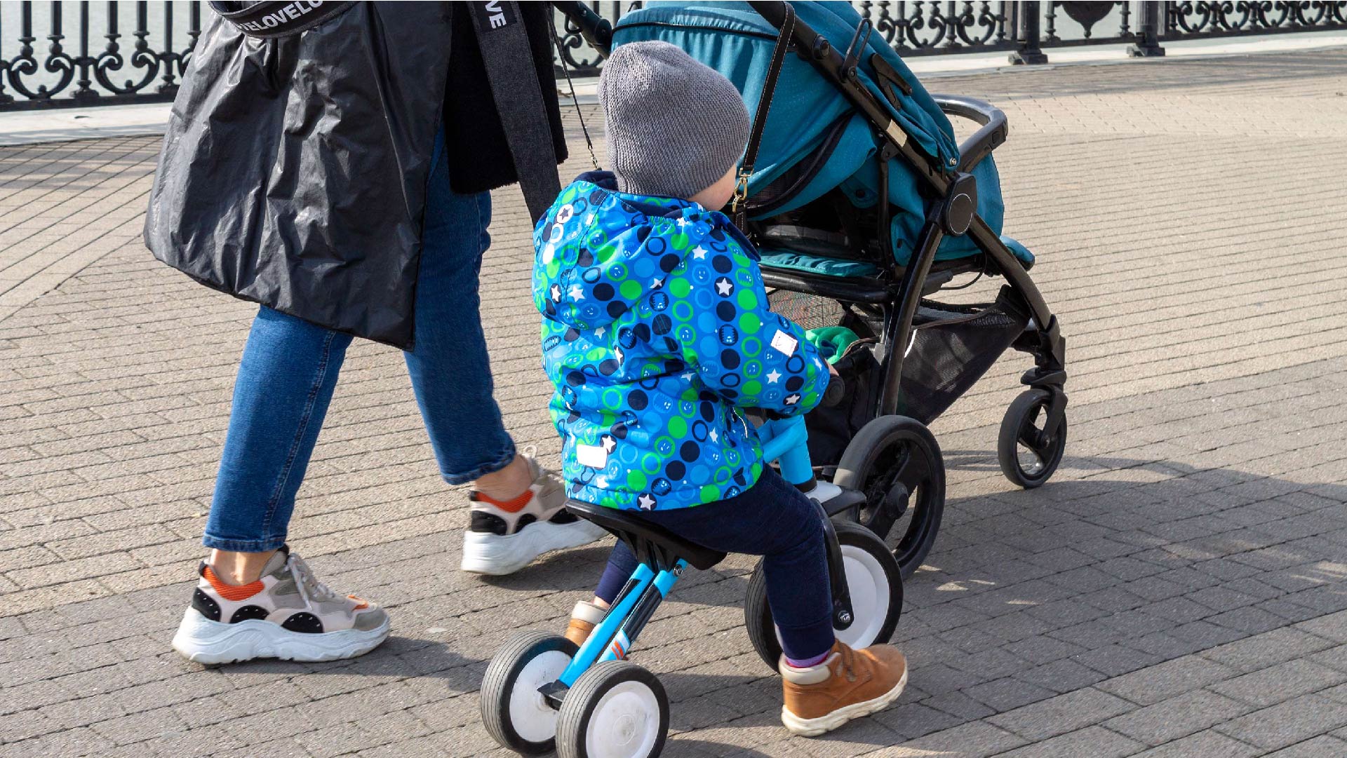 Een mama duwt de kinderkoets met haar zoontje die haar op een driewieler volgt